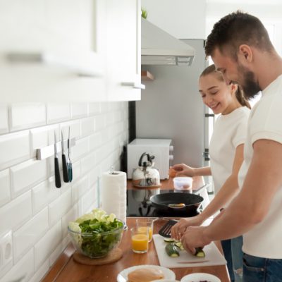 Happy young couple cooking breakfast in modern kitchen together, smiling wife making omelette or scrambles eggs, husband slice vegetables, lovers prepare food spending morning at home